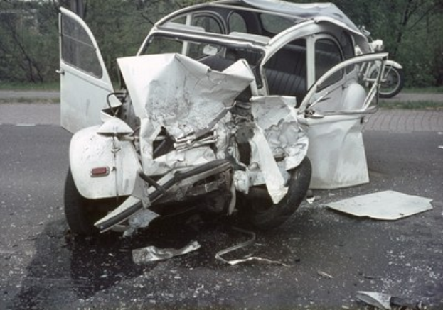 Schwer beschädigte Frontpartie eines Autos am Stra√enrand mit Bäumen und einem Gebäude im Hintergrund, ein weiteres Fahrzeug in der Ferne sichtbar.