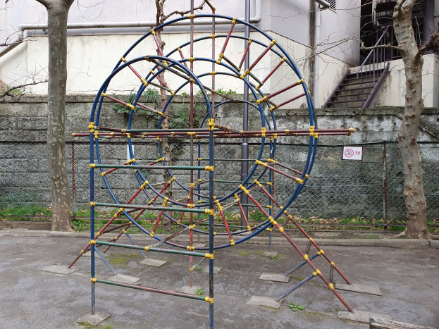 Spielplatz mit großem Metallgerüst, umgeben von Bäumen und einer Maschendrahtzaun, mit Treppen zu einem Gebäude mit Fenstern und Rohren, einem Schild an einem Pfahl und grasbewachsenem Boden.