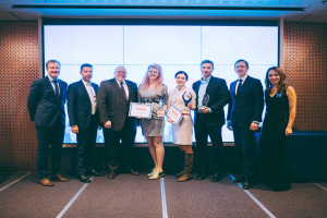 Gruppe strahlender Menschen mit Urkunden, die vor einem Bildschirm mit einem schwarzen Tischtuch und einem Wandobjekt im Hintergrund stehen, Gewinner der International Business Awards 2018