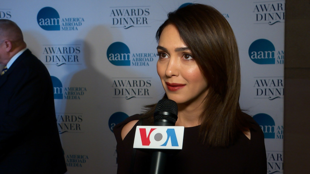 Emmy Rossum in einem schwarzen Kleid mit einem Mikrofon in der Hand, mit einem Mann links von ihr und einem Banner mit der Aufschrift "Aam Awards Dinner" im Hintergrund.