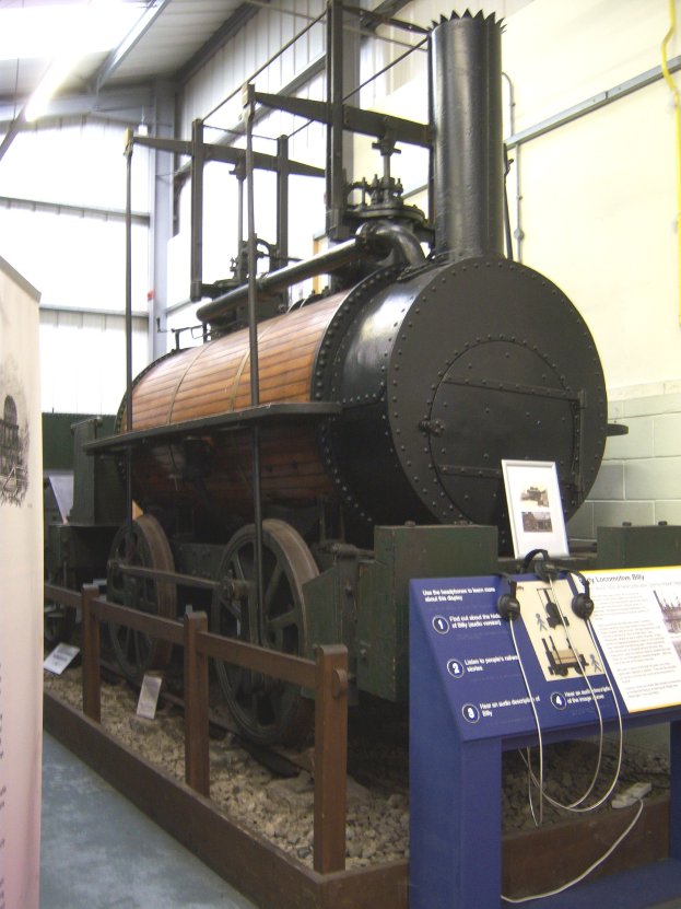 Alte Dampflokomotive in einem Museum ausgestellt, umgeben von Informationsschildern, einem Zaun, Steinen, einer Fahne, einer Wand mit Rohren und Deckenlampen.