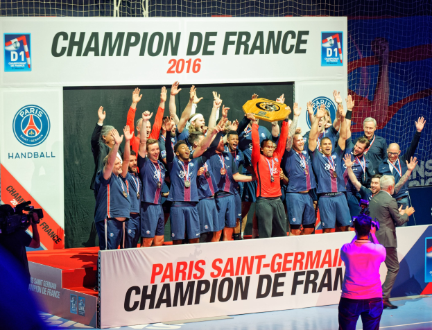 Eine Gruppe von Männern auf einer Bühne mit einem Pokal, wobei eine Person den Moment mit einer Kamera aufnimmt und ein "Champion de France 2016"-Schild im Hintergrund zu sehen ist.