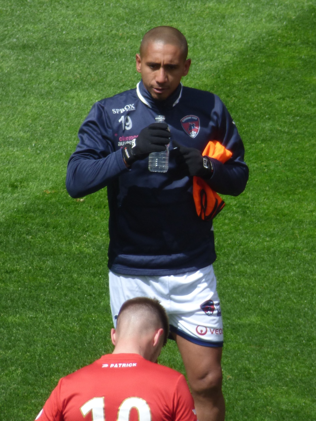 Ein professioneller Athlet steht auf einem saftig grünen Feld und hält in einer Hand eine Flasche und in der anderen ein oranges Tuch.