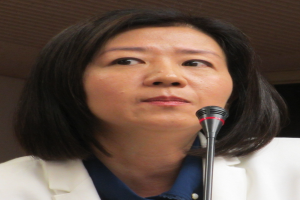 Eine Frau in einer weißen Jacke und einem Anzug spricht in ein Mikrofon vor einer Wand und sieht ernst aus.