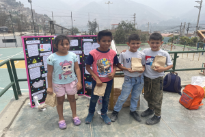 Gruppe von Kindern auf einem Basketballfeld mit Papiertüten in den Händen, mit Vorräten auf dem Boden, einer Tafel mit Text links daneben und einem Hintergrund aus Zaun, Pfosten, Straßenlaternen, Bäumen, Gebäuden, Hügeln und Himmel.