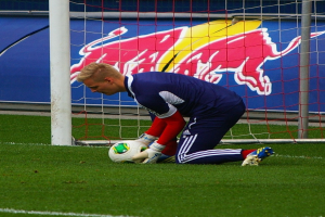 Ein Fußballspieler kniet vor einem Tor mit einem Ball auf dem Boden, umgeben von Gras und einem Netz, mit einer Fahne mit Text und einem Bild einer Person im Hintergrund.