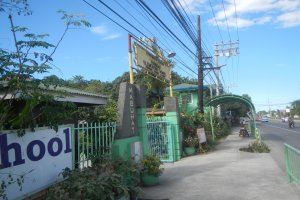 Schuleingang mit einem "Schule"-Schild am Straßenrand, fahrende Fahrzeuge, ein Tor, ein Tor, eine Namensschild, Pflanzen, Bäume, Strommasten mit Drähten und ein bewölkter Himmel im Hintergrund.