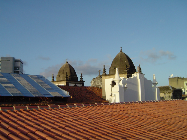 Stadtansicht mit Gebäuden im Vordergrund, Solarpanelen auf einem Dach und einem blauen Himmel im Hintergrund.