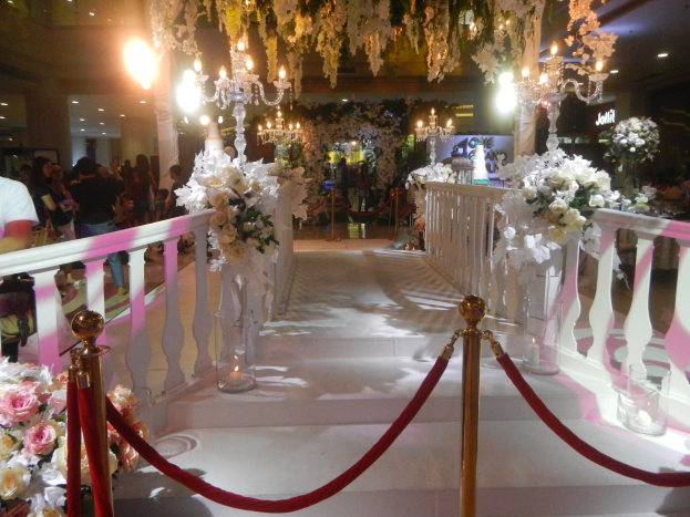 Eine Trauung in einem Einkaufszentrum mit weißem Teppich, dekorativen Geländern, Blumenarrangements und Deckenbeleuchtung, bei der eine Menge Menschen mit Taschen anwesend sind.