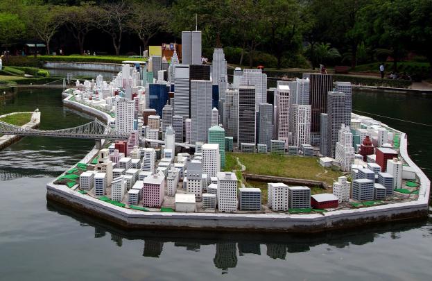 Ein detailliertes Miniaturmodell einer Stadtlandschaft mit Gebäuden, Grünflächen, einer Brücke und Menschen, das in einem Gewässer gegen einen Himmel als Hintergrund gesetzt ist.