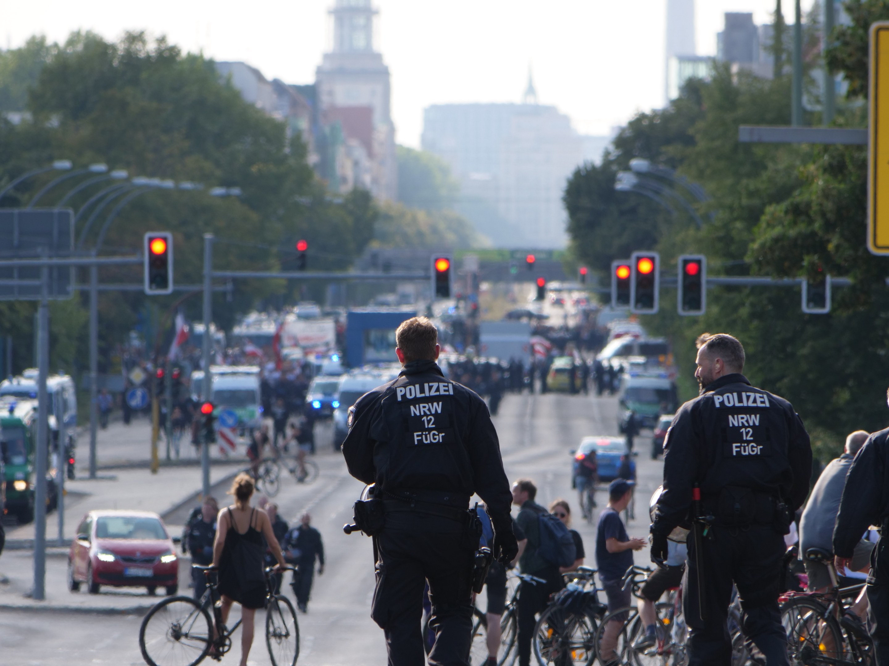 Polizeibeamte fahren auf Fahrrädern eine von Bäumen gesäumte Straße mit Gebäuden und einem klaren blauen Himmel im Hintergrund.