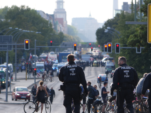 Polizeibeamte fahren auf Fahrrädern eine von Bäumen gesäumte Straße mit Gebäuden und einem klaren blauen Himmel im Hintergrund.