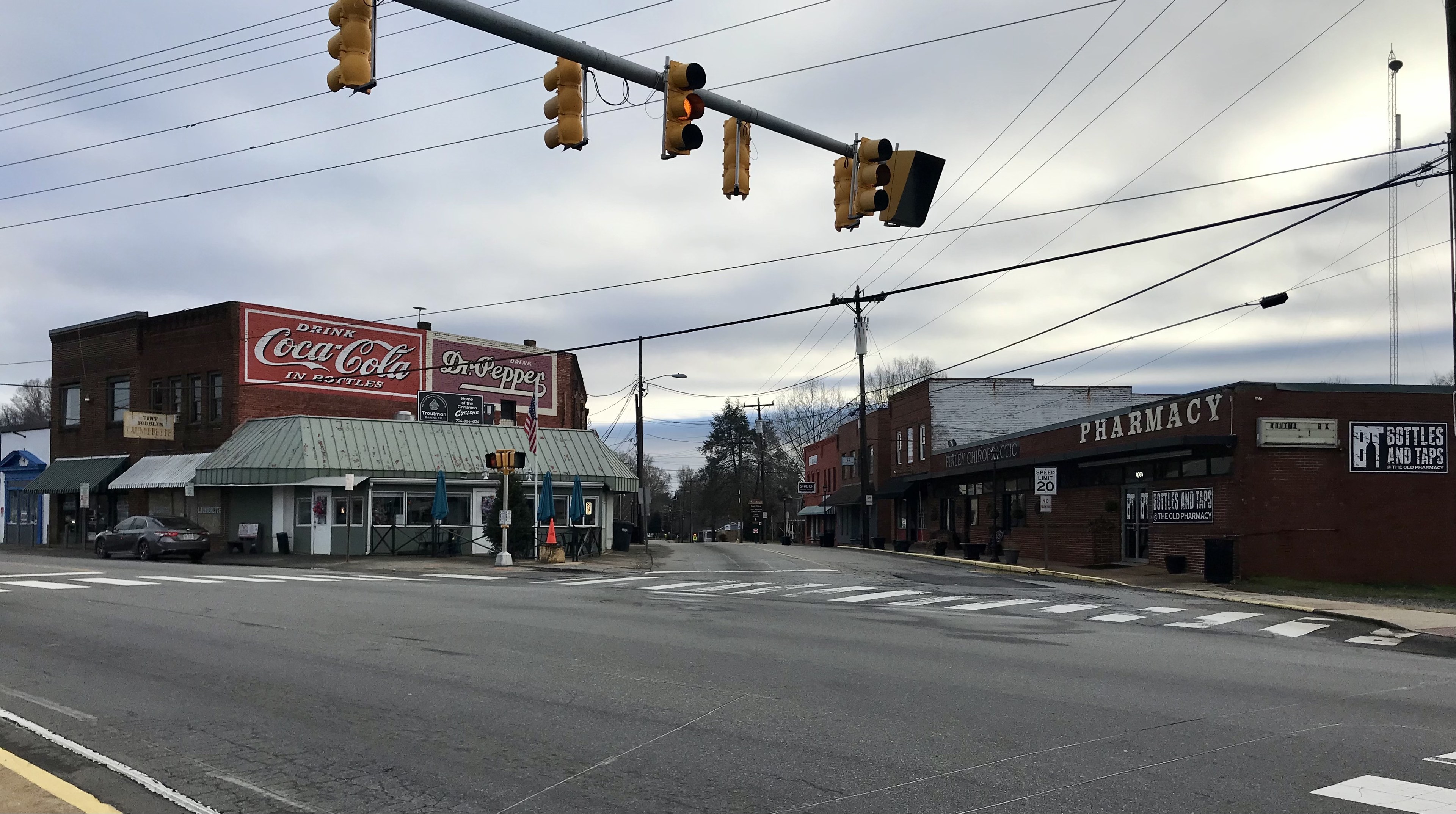 Stadtstraße mit einer Ampel an einer Kreuzung, Fahrzeuge, Gebäude mit Beschriftung, Strommäste mit Drähten, Bäume und ein bewölkter Himmel.