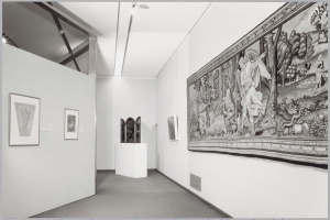 Schwarzes und weißes Foto einer Museumsgalerie mit einem großen Wandteppich, gerahmten Kunstwerken an den Wänden, auf einem Sockel montierten Objekten und Deckenleuchten, mit "Museum of Fine Arts, New York City" unten sichtbar.