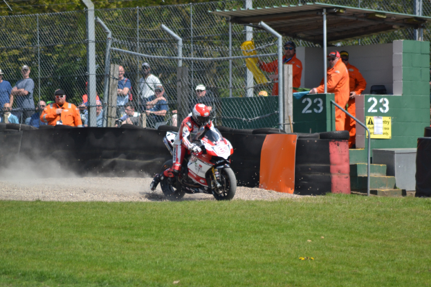 Ein Motorradfahrer in Helm und Handschuhen fährt über ein grasbewachsenes Feld, mit Zuschauern in Kappen und Schutzbrillen, einem eingezäunten Bereich, Treppen mit Geländern und einem nummerierten Schuppen im Hintergrund.