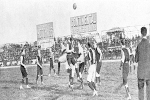 Ein Schwarz-Weiß-Bild einer Gruppe von Männern, die auf einem Feld Fußball spielen, mit einem Ball in der Luft und einer Zuschauermenge im Hintergrund, mit Tafeln, die wahrscheinlich textbezogen zum Spiel sind.