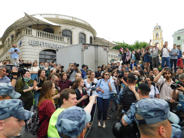Große Menschenmenge vor einem Gebäude protestierend, einige mit Kameras und Handys, mit einem Fahrzeug, Bäumen und klarem blauem Himmel im Hintergrund.