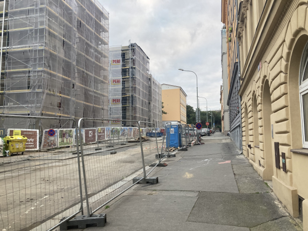 Stadtstraße mit Gebäuden, Fahrzeugen, Straßeninfrastruktur, Bäumen und einer Baustelle mit Gerüst im Vordergrund.