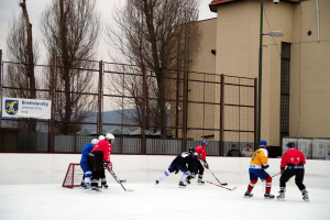 Menschen, die Eis-hockey auf einer Eisbahn mit Geb√§uden, B√§umen, einer Stra√Ÿenlaterne, einer Namens-plakette und Z√§unen im Hintergrund unter einem klaren Himmel spielen.