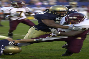 Zwei Fußballspieler in Helmen und Uniformen kämpfen auf dem Feld, während andere im Hintergrund zusehen.