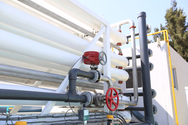 Große Gruppe von Rohren, Ventilen, Metallstangen und anderen Komponenten auf einem Gebäudedach mit Bäumen und einem klaren blauen Himmel im Hintergrund.