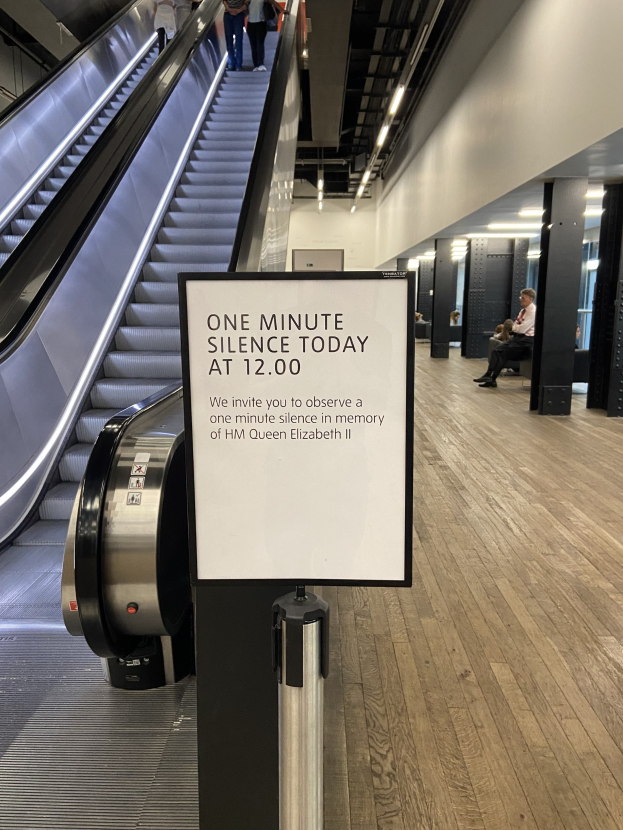 Eine Rolltreppe im Flughafen mit einem Schild, auf dem "Eine Minute Stille heute" steht, ein paar Menschen darauf und in der Hintergrundbeleuchtung an der Decke befestigte Lampen.