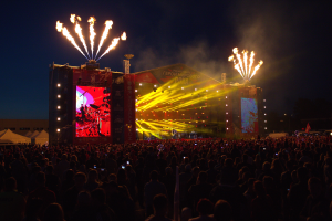 Eine große Menschenmenge auf einem Musikfestival mit einer hell erleuchteten Bühne mit Performern und Bannern, Zelte links und ein Feuerwerk, das den dunklen Himmel erhellt.