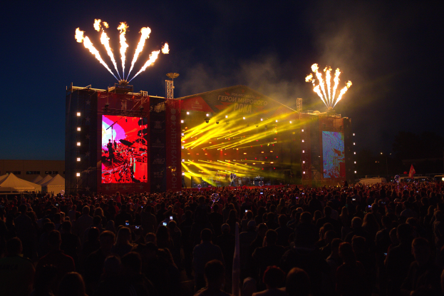 Eine große Menschenmenge auf einem Musikfestival mit einer hell erleuchteten Bühne mit Performern und Bannern, Zelte links und ein Feuerwerk, das den dunklen Himmel erhellt.