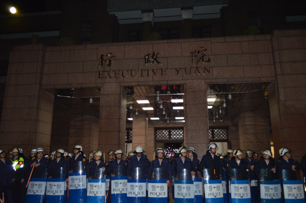 Eine Gruppe von Polizisten in Helmen und Schutzausrüstung steht vor einem Gebäude mit beleuchteter Schrift, haltend Schilde mit Text, mit zusätzlichen Gebäuden im Hintergrund.