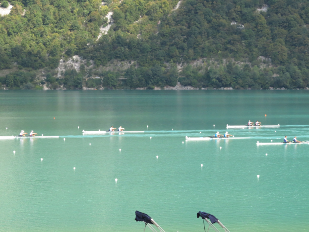 Gruppe von Menschen beim Ruderwettbewerb auf einem Gewässer mit Bäumen und einem Hügel im Hintergrund, Kleidung über Stühle drapiert.