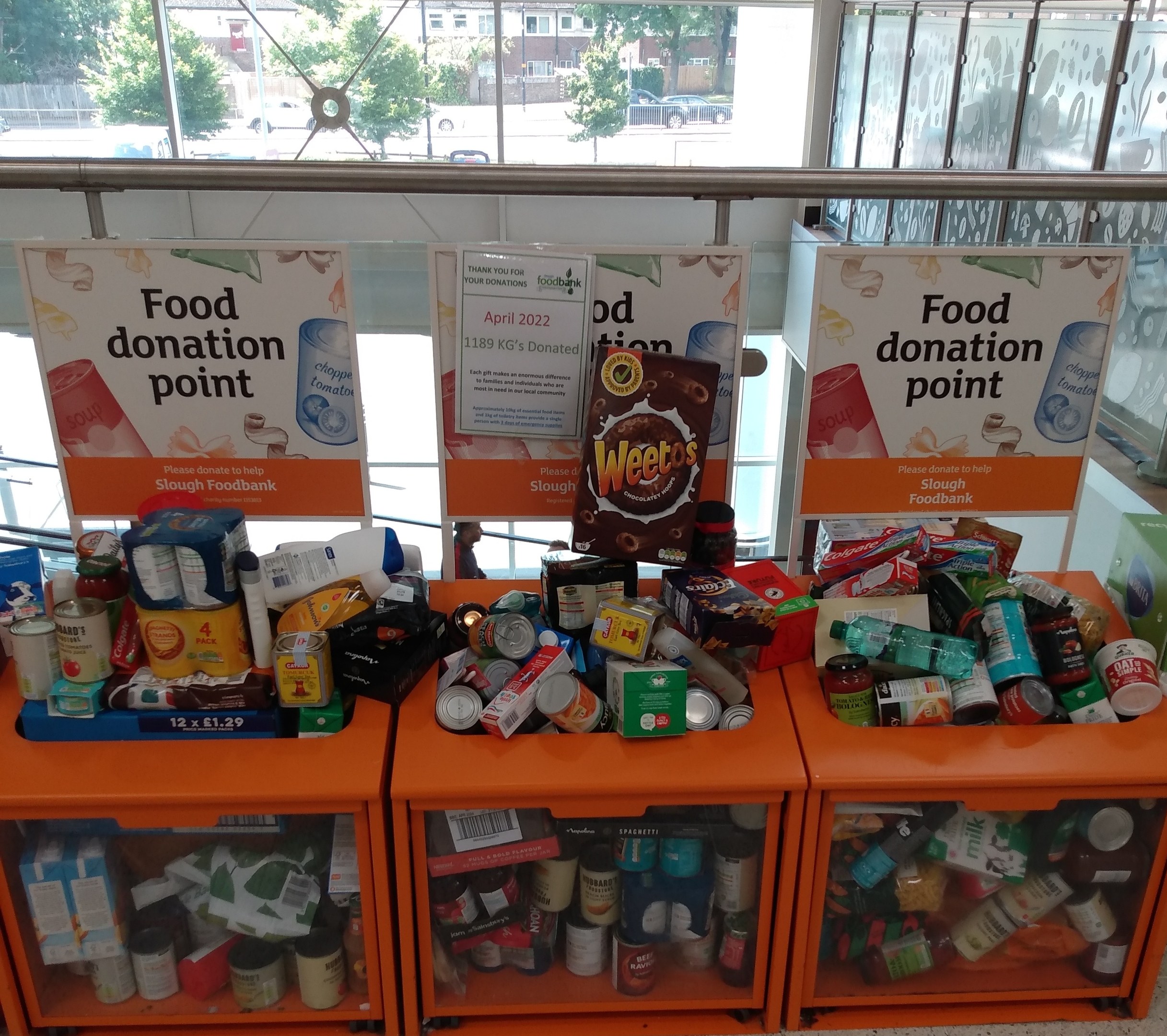 Orange Schränke mit Lebensmittelspenden gefüllt, darunter Flaschen, Dosen und Kartons, mit Informationsschildern vor einem Hintergrund aus Fahrzeugen, Bäumen, Gebäuden und Himmel durch Glasfenster sichtbar.