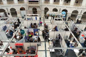 Eine große Gruppe von Menschen versammelt sich in einem Raum mit Ständen, Bannern, Tischen, Stühlen, Luftballons und einem Gebäude mit Bögen, Säulen, Geländern und einem Bildschirm im Hintergrund.