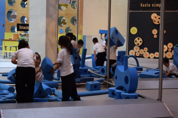 Kinder beim Spielen in einer Turnhalle, einige halten Gegenstände, mit einer Texttafel an der Wand und Stangen rechts.