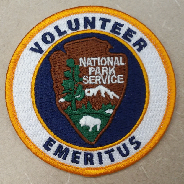 Runder Aufnäher mit blauem Rand und weißem Hintergrund, der den schwarzen Text "Volunteer National Park Service Emeritus" in der Mitte eines gelben Kreises zeigt.
