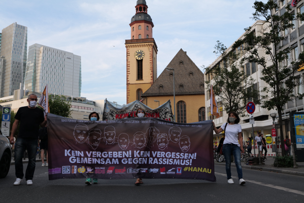 Eine Gruppe von Menschen mit Masken, die eine Straße entlanggehen und ein Transparent halten, mit einem geparkten Auto auf der linken Seite, Gebäuden, Bäumen, Schildern, Pfählen, einem Kirchturm und einem klaren blauen Himmel im Hintergrund.