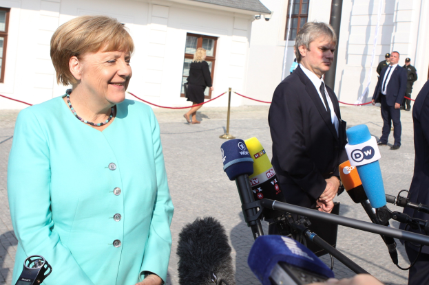 Bundeskanzlerin Angela Merkel spricht vor einem Gebäude mit Fenstern zu Reportern, umgeben von Menschen und Medienpersonal mit Mikrofonen, unter einem bewölkten Himmel.