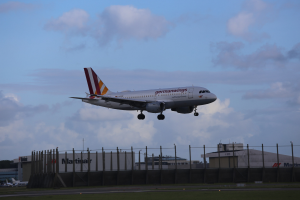 Germanwings Airbus A320-200 beim Start von Frankfurt Airport mit Gras darunter, einem Zaun im Vordergrund, Gebäuden und Bäumen im Hintergrund und einer bewölkten Himmel.