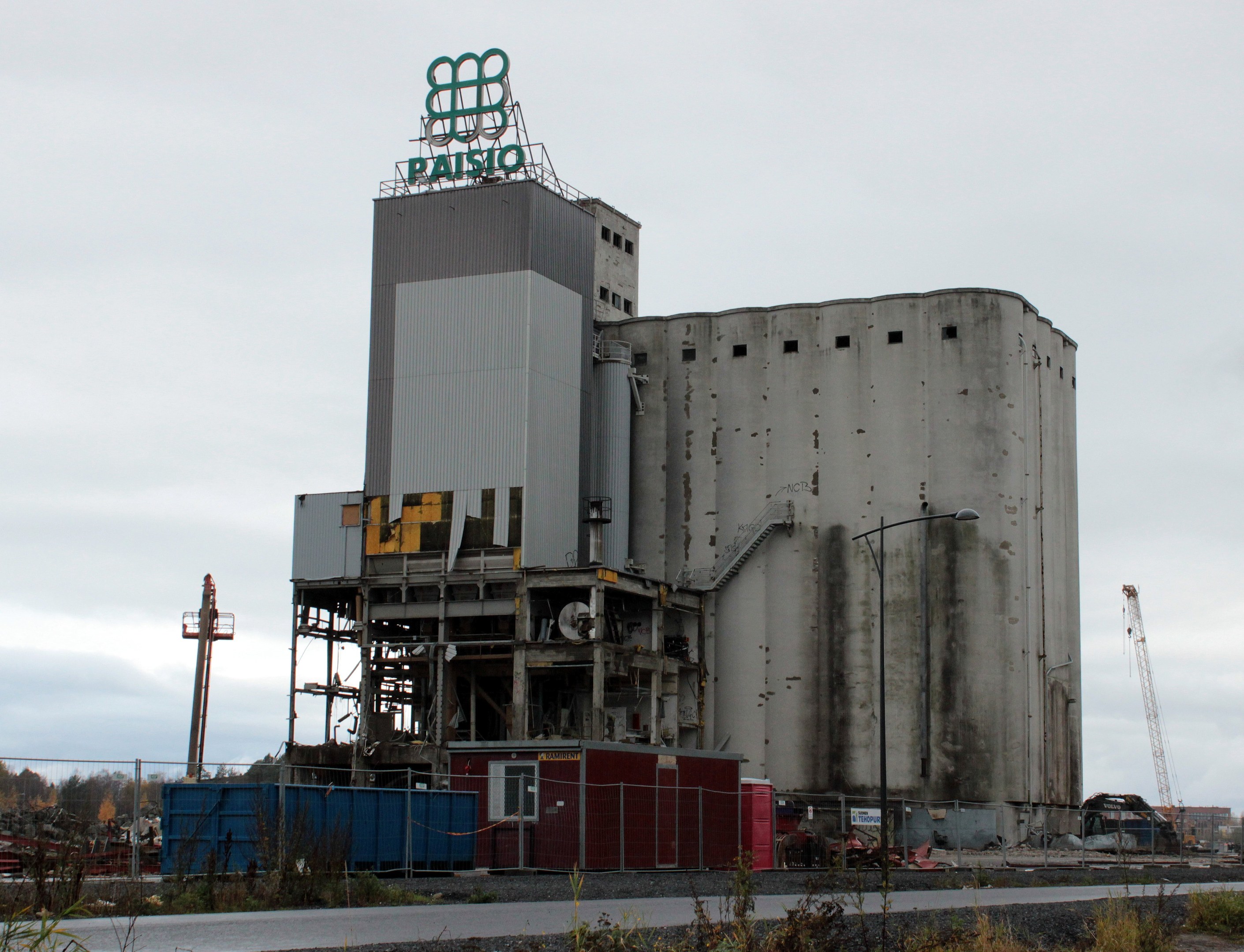 Großes Zementgebäude neben einer Straße, umgeben von Pflanzen, einem Zaun, Pfählen, Lampen und anderen Gegenständen, mit Bäumen und einem klaren blauen Himmel im Hintergrund.