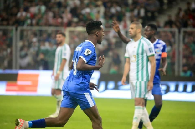 Fußballspieler in einem blauen und weißen Trikot, der nach einem Tor vor einer jubelnden Menge feiert, umgeben von Teamkollegen in weißen und grünen Trikots, mit einem weißen und blauen Brett und Geländer im unscharfen Hintergrund.