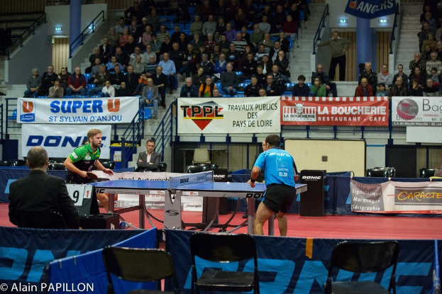 Indoor-Tischtennis-Match mit spielenden Spielern, sitzenden Zuschauern und einigen stehenden Beobachtern sowie leeren Stühlen und Werbetafeln.