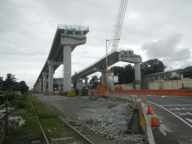 Baustelle mit einer Brücke im Hintergrund, Straße durch Verkehrskegel markiert, Eisenbahnschiene links, verstreute Steine und Gras, Bäume und Gebäude säumen die Straße und ein bewölkter Himmel.
