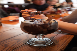 Eine Gruppe von Menschen sitzt um einen Tisch, in der Mitte ein Glas Schokoladenpudding, eine Person im Vordergrund isst mit einem Löffel, Hintergrund leicht unscharf.