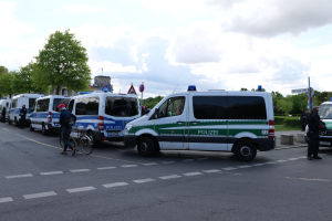 Eine Gruppe von Polizeiwagen entlang einer Straße mit einem Radfahrer im Vordergrund, Bäume, Masten, Schilder, Gebäude und ein bewölktes Himmel im Hintergrund.