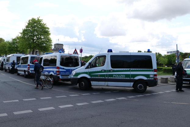 Eine Gruppe von Polizeiwagen entlang einer Straße mit einem Radfahrer im Vordergrund, Bäume, Masten, Schilder, Gebäude und ein bewölktes Himmel im Hintergrund.