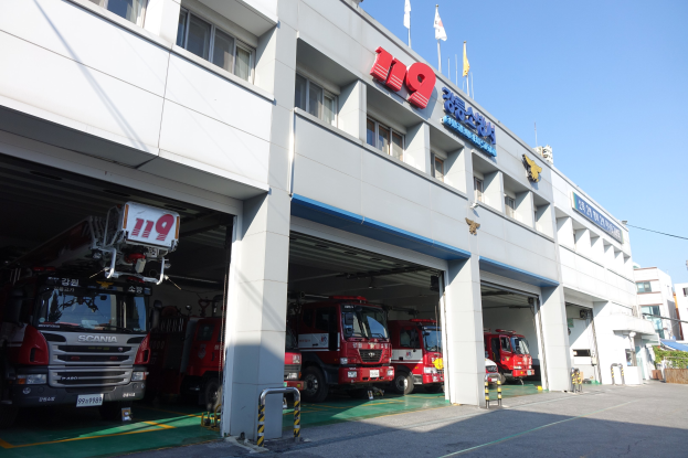 Feuerwache mit mehreren Feuerwehren vor dem Gebäude geparkt, mit Fenstern, Säulen, Namensschildern und Fahnenmasten im Vordergrund, vor anderen Gebäuden und einem klaren blauen Himmel.
