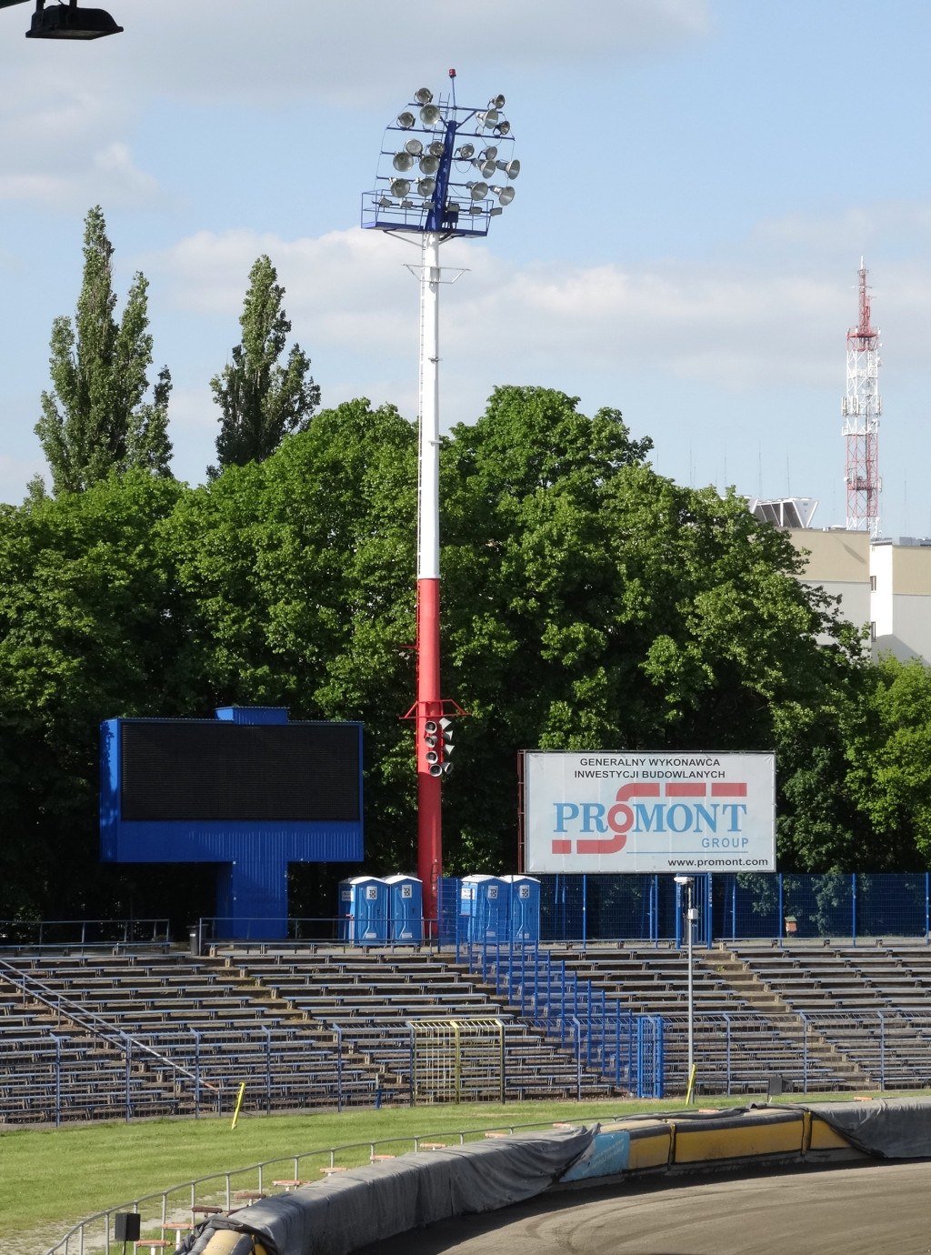 Rennstrecke mit Flutlicht, umgeben von Gras, Geländern, Stufen, einem Lichtmast, einem Schild, Bäumen, Gebäuden, Türmen und einem bewölkten Himmel.