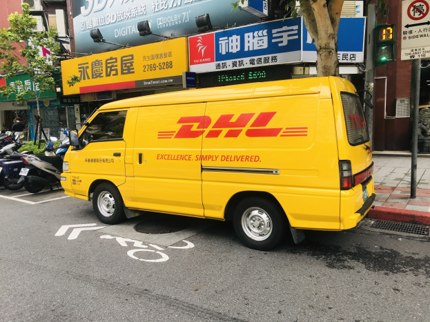 Gelbes DHL-Lieferfahrzeug auf der Straßenseite mit Gebäuden, Bäumen, Schildern, Masten, Ampeln und einer Person auf dem Gehweg.