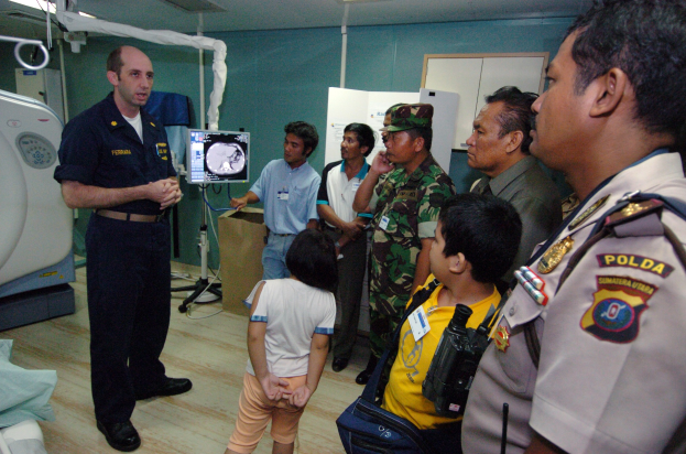Eine Gruppe von Menschen steht in einem Krankenhauszimmer, ein Mann hält ein Mikrofon in der Mitte, ein Bett mit einer Decke darauf links, verschiedene medizinische Geräte, ein Röntgengerät, eine Tafel mit Text, eine Tür, eine Wand und eine Decke mit Lampen im Hintergrund.