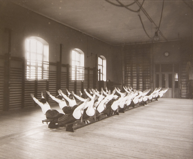 Gruppe von Menschen, die Pilates-Übungen in einem Fitnessstudio mit Fenstern und einer Uhr an der Wand machen.