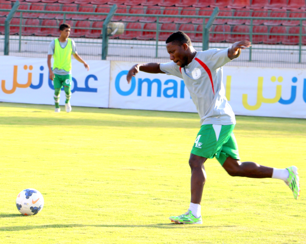 Ein Mann kickt einen Fußball auf einem Rasenfeld in einem Stadion, umgeben von zwei Personen und Bannern, Geländern und Stühlen im Hintergrund.